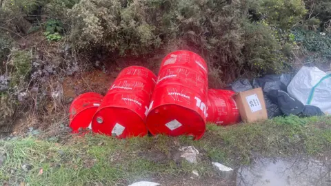 Buckinghamshire Council Four red barrels are in a ditch with other discarded boxes and bags.