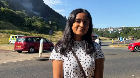 PA Media Mahek Jayantilial stands in front Arthur's Seat. She has dark hair and is wearing a black and white spotted t-shirt. 