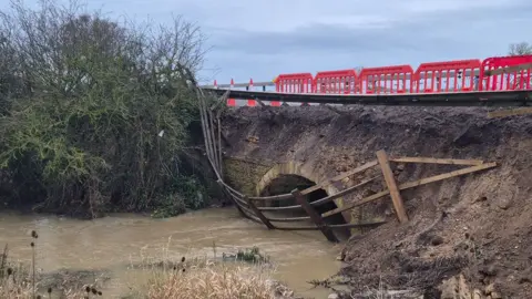 A damaged culvert under the A1 southbound over Billing Brook, Water Newton. It shows a damaged traffic barrier tipping over a muddy bank leading to the brick-built arched culvert opening. A wooden fence has tumbled down the bank and is tipped into the water, which is muddy with silt. There are shrubs to the left of the brook. A red crash barrier and red and white cones run along the edge of the road above the culvert. 