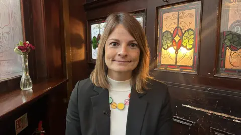 Janine Hirst wearing a white rollneck jumper, black blazer and a brightly-coloured necklace is picture in front of a dark wood background that has stained glass patterned windows on the top