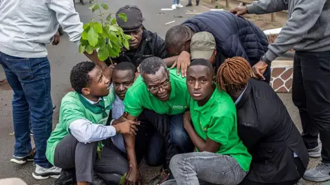 Eight people crouched in a huddle and they are being detained. There is a person standing on either side, keeping them down