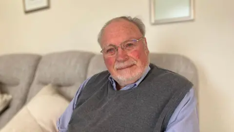 BBC/Sam Dixon-French David Barker, wearing a light purple shirt and a grey vest, sitting on a white sofa inside his living room.
