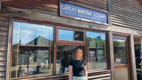 A woman in a black t-shirt and green and white striped trousers, stands outside a wooden framed shop. A sign across the top says Great British Jigsaws
