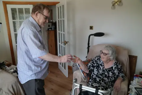 A photo of David and Anne Summers in their house in Llandrindod Wells. David is giving Anne a cup of tablets that she needs to take. They are in the living room with a sofa and seat in the picture.