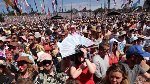 Woman with fan over her head, stood next to people with sun hats and sunglasses