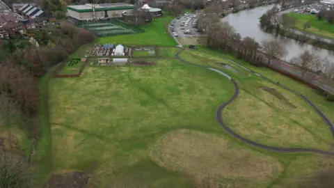BBC an aerial shot taken by a drone, of a green field crisscrossed by paths, on which some polytunnels are visible. There is a bit sports building in the background, and running alongside the field are a road and a river