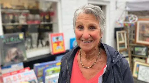 A woman with grey hair, wearing a dark blue jacket over a pink top and necklace.
She is smiling, and there are games and pictures displayed behind her 