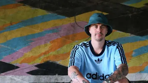 Rikki, a man, wearing a blue adidas jersey and black shorts and a round blue hat and purple shoes, squatting on the ground covered with graffiti.