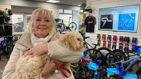 A woman wearing a white coat and scarf and holding a small white dog. A row of bikes with blue signs and bows on them are behind her. She is in a cycle shop. The floor is wood and there are white walls with cycle equipment around. 