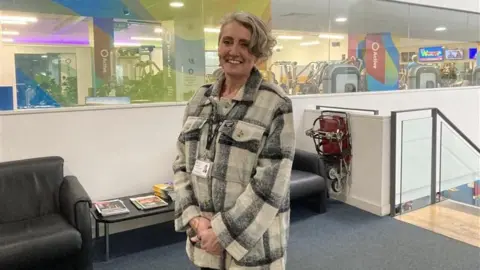 A woman with short fair hair, and a black and grey checked jacket smiles in front of the camera