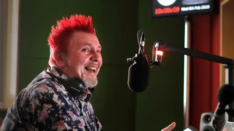 Teesside Live Dave Llewellyn sitting in a radio studio in front of a microphone. He is wearing a dark shirt with a pink floral pattern and has spiked red hair. He has a grey goatee and is laughing.