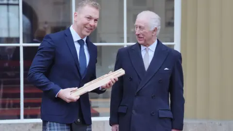 Sir Chris Hoy holds the baton as King Charles stands next to him withing the grounds of Buckingham Palace. The King is wearing a blue coat with a light shirt and grey tie. Sir Chris is wearing a blue jacket and kilt, a white shirt and a blue tie