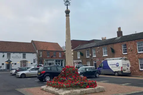 Jake Zuckerman/BBC A wider view of the monument, which now stands in the middle of a carpark in the centre of the town.  Old buildings provide a backdrop to the scene.