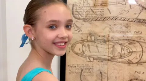 Robby West/BBC Jessica is wearing a blue leotard and is standing in front of a drawn diagram of a ballet shoe, which is stuck on a white wall. She is turn her head over her shoulder, smiling, looking at the camera.