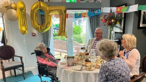 Ms Jones sitting at a table where a high tea has been set. There are three people sitting with her, and in the background there are balloons and lots of cards hanging on a string