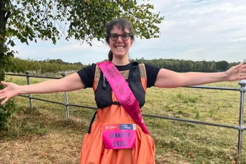 Supplied Jennifer Gane is standing in a field and is wearing an orange ballgown and a pink sash. She also has a Great North Run competitor number pinned to her front which reads: "Jennifer 54351". She is smiling broadly and has her arms outstretched.