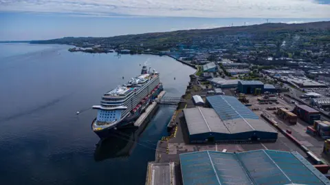 Getty Images An image of Greenock harbour, with a large cruise ship docked in the foreground, next to a shore studded with industrial buildings. Wind turbines are visible on the hills of the horizon, while the water reflects the clouds in the sky