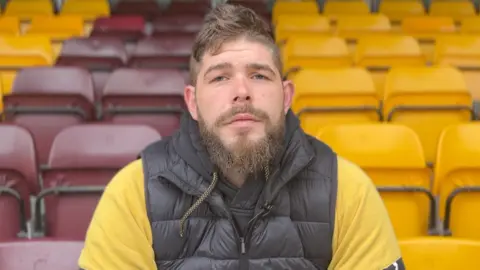 James Vincent/BBC A man with brown hair and a beard sits with empty rugby stadium seats behind him. He is wearing a black sleeveless jacket and yellow top underneath.