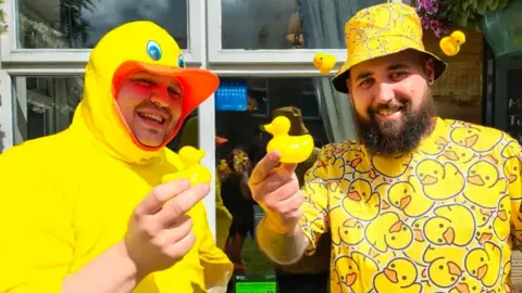 Daniel Caldwell dressed as a duck in a bright yellow hoodie with a duck's beak open over his face and Curtis Lunt in a yellow bucket hat and tshirt covered in yellow ducks, both smiling and holding rubber ducks