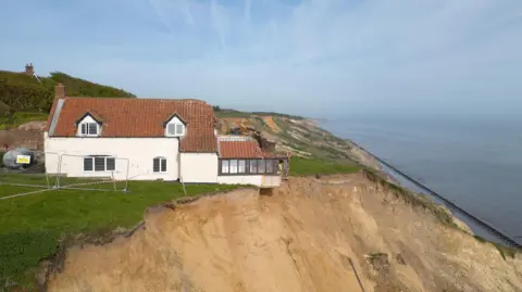 A white farmhouse with a tiled roof teetering on the edge of a sandy cliff. The farmhouse is surrounded by grass and a metal fence has been erected around the property. The sea and Norfolk coastline can be seen in the background