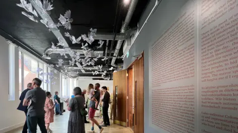 People come into the exhibition and look around. There is writing on the wall describing the internment camp and there are paper dovers hanging from the ceiling.