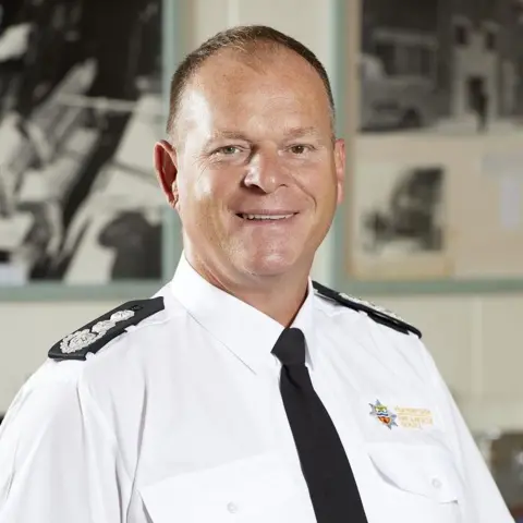 Humberside Fire and Rescue A portrait photograph of chief fire officer Phil Shillito. He has short cropped hair and is smiling into the camera. He is wearing a white shirt with a Humberside Fire and Rescue logo, black lapel badges and a black tie.