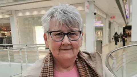 A woman stands in a shopping mall. she is wearing n=a pink jumper, checked scarf and grey coats. she has short grey hair and is wearing glasses.