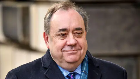 Getty Images A smiling man with receding hair is wearing a dark coat, tartan scart, blue shirt and tie