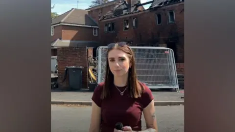 Millie Trenholm is stood outside the affected properties. She has long brown hair with her sunglasses on her head. She is wearing a dark red t-shirt and is clutching a black microphone. 