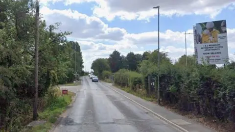 Google A Google Maps photo of a narrow road with trees either side of it. A turning can be seen to the left with cars in the distance and a sign on the right which says "bedroom retirement living apartments".