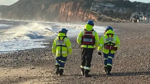 BBC Three emergency service workers dressed in hi-visibility clothing and blue hard hats walking on a stone beach. They are wearing fluorescent clothing. A clifftop is seen in the background. There are large waves breaking onto the shore.