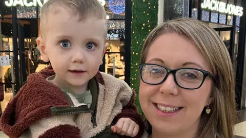 A woman with blonde hair, a white top and glasses is holding a young child inside a shopping centre. The child has blonde hair and is wearing a multi coloured top.
