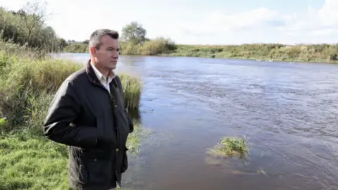 A man with short dark hair and a green waxed jacket looking out across a small river with grassy banks on either side