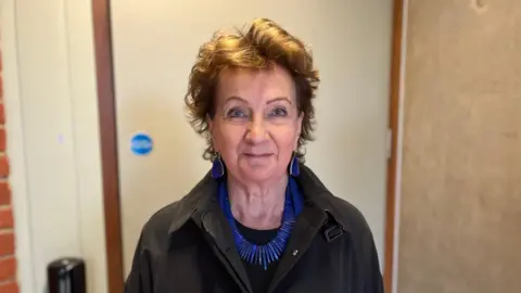 Roisín McDonough wearing a black coat with blue jewellery, she has brown hair 