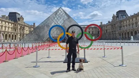 West Midlands Police A police officer in all black clothes holds the lead of a brown and white dog, who is sat on the ground, in front of a statue of the five Olympic rings and the Louvre