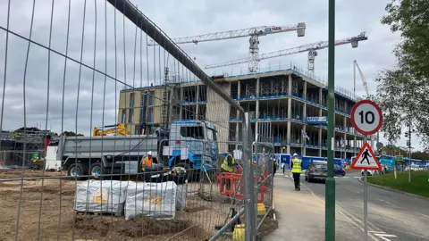 Building work at the Royal Shrewsbury Hospital. A number of people in hard hats are doing building work. A large crane towers over a partially built building, with a wire fence in the foreground