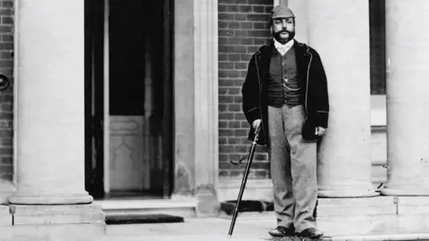 A black and white photo of Maharajah Duleep Singh with walking stick outside Elveden Hall. He is standing beside a column towards the entrance to the hall. 
