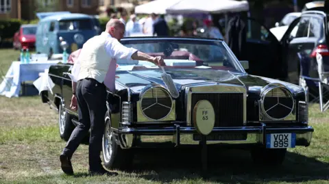 Reuters A racegoer cleans their car ahead of the day's race. He is dressed in a suit and is using a soft brush to clean one of its headlights.
