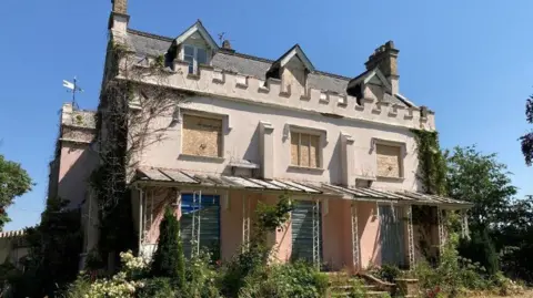 A large pink building which is all boarded up sits in green grass underneath a blue sky. 
