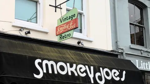 A Regency terrace, comprised of one cream coloured building and a pale grey one. There is a large black awning with the words "Smokey Joe's" in white writing and a sign above it that reads "Vintage Smokey Joe's Retro".