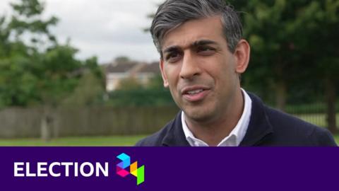 Rishi Sunak wearing a navy blue zip-up top and white shirt looks off camera, as he stands outside in a park with trees behind him. The words election 24 are overlaid on the image