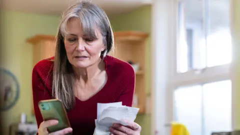 Getty Images Middle-aged woman with long grey hair looks at a pile of receipts and her mobile phone. She has a slightly anxious look on her face.