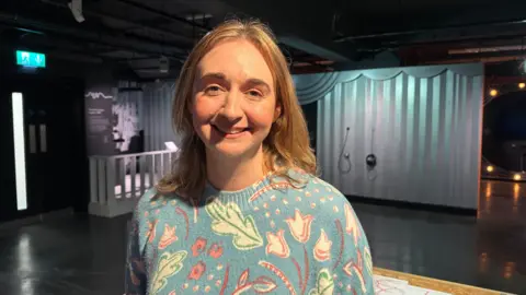 Jessica Bayley/BBC A woman wearing a blue jumper with flowers on it smiles at the camera, she has shoulder length blonde hair and behind her you can see part of an exhibition hall.