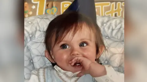 Lucy Owen A baby smiling, he has his left hand in his mouth and is wearing a blue party hat