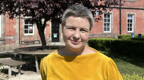 Yorkshire Cancer Research A woman in a mustard shirt stands in front of a red brick building with a tree and a bench.