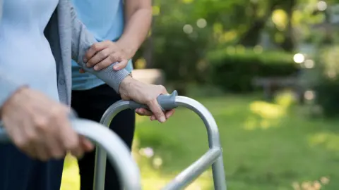 An older person has their hands on a walking frame and are being helped by a care giver.