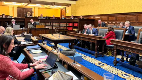 Members sitting in the Tynwald Chamber, which has wooden wall panels and benches with blue leather-clad seats and a blue carpet.