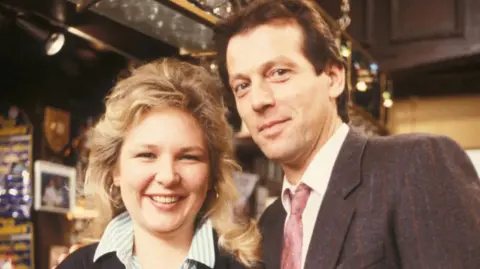 Kathryn Apanowicz is smiling as she stands alongside Leslie Grantham playing Den Watts in the Queen Vic pub.