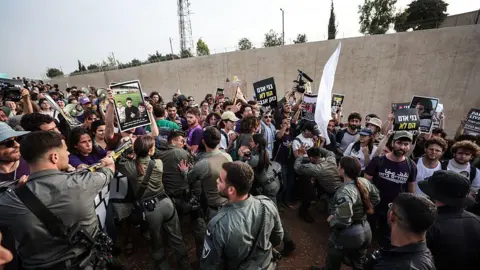 Anti-war protesters, carrying signs, are held back by security forces.
