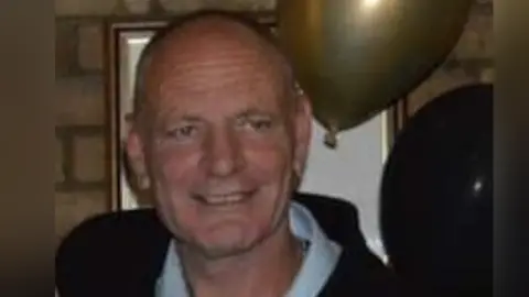 Gary wearing a baby blue shirt and a black jacket. He is at a party with balloons in the background and he is smiling at the camera.